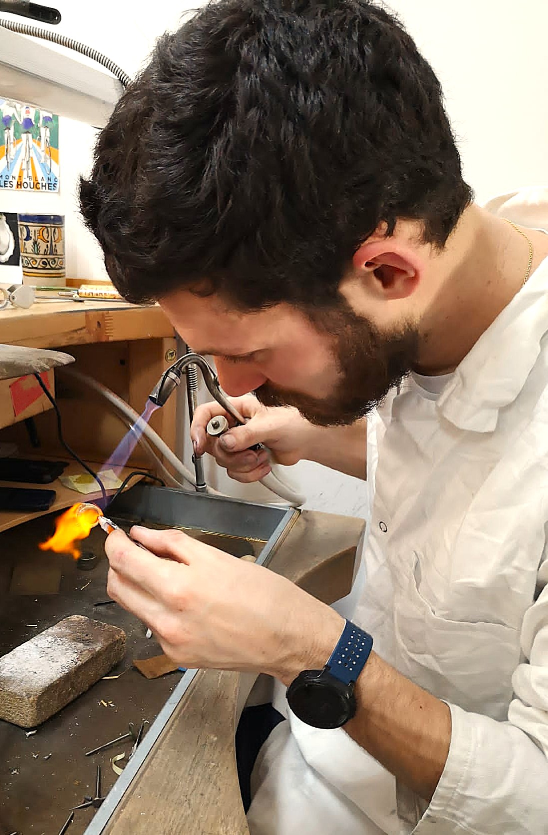 L'image représente un artisan bijoutier joaillier au travail sur son établi. Il est en train de souder un bijou en or à la flamme. C'est un travail traditionnel.